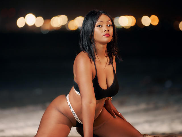 MayaRichh has dark skin, black hair, and is wearing a black sports bra and white Calvin Klein thong, kneeling on a beach at night with bokeh lights.