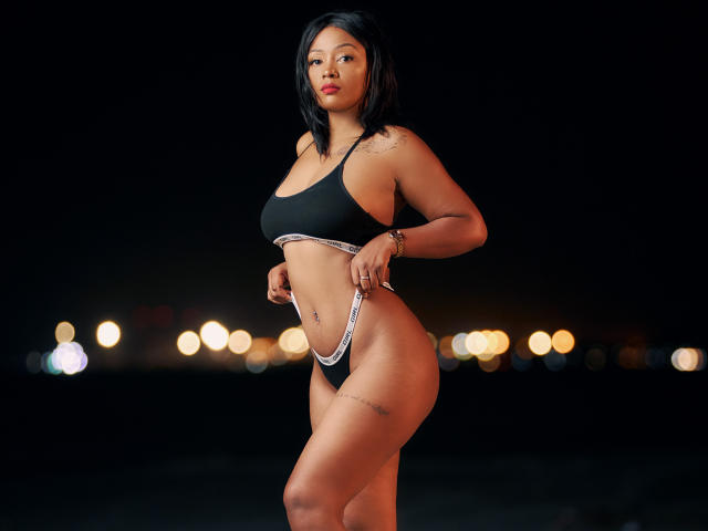 MayaRichh with medium brown skin, black hair, red lipstick, wearing a black sports bra and white-trimmed black thong, standing, showing her curvy ass and small tattoos on her thigh.
