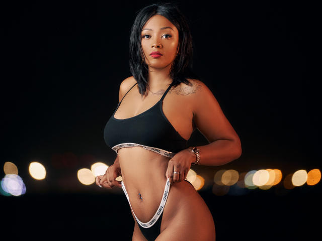 MayaRichh with black hair, wearing a black sports bra and white Calvin Klein thong, standing, looking at camera, showing her small ass and medium tits, night background.