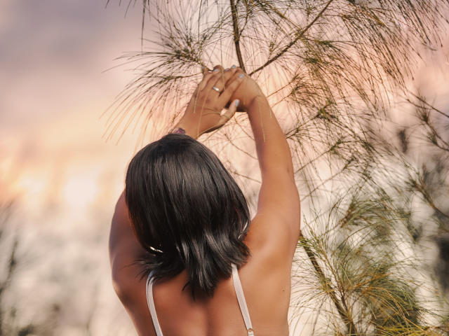 MayaRichh with dark, shoulder-length hair, arms raised, touching thin, green, grass-like branches, wearing a white, sleeveless top, sunlight highlighting her back.