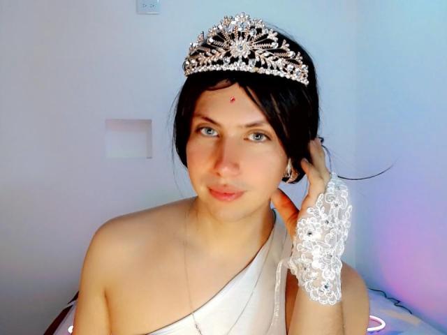 AdriudI with dark hair, wearing a silver tiara, white lace glove, and white sash, has a red dot on her forehead, and is smiling with blue eyes.