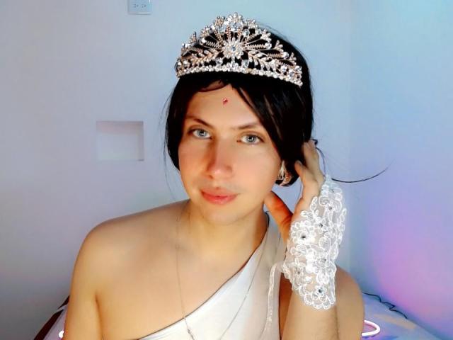 AdriudI with a silver tiara, wearing a white one-shoulder dress and a lace glove, touching their black hair, has blue eyes and a red dot on their forehead.