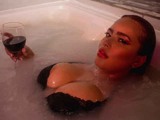 LaurenMartins in a bathtub, holding a glass of red wine, with her big tits and wet black lace bra, looking sexy with red lipstick and slicked-back brown hair.