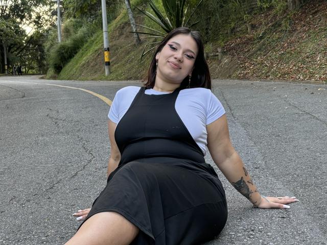 Milly Johnson, con un seno di media grandezza, indossa un abito nero con maniche bianche, seduta su una strada, con le braccia dietro la schiena, sorridente, con un tatuaggio sull'avambraccio destro, capelli scuri, pelle chiara, sfondo verde, strada, giorno..