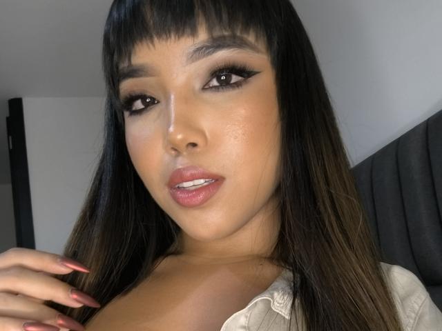 Heyly with long black hair and heavy eyeliner, giving a sultry look while her hand with red nails rests on her chest.