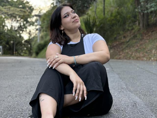 MillyJohnson with dark brown hair, white t-shirt, black dress, and white nails, sitting on a road, arms crossed, legs bent, looking up with a slight smile, tattoos visible.