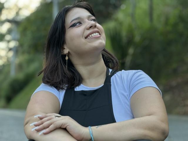 Milly Johnson, com seios médios, sorrindo, braços cruzados, vestindo um avental preto sobre uma camisa branca, cabelo castanho escuro, delineador preto, brincos dourados, pulseira azul, ao ar livre, fundo verde desfocado..