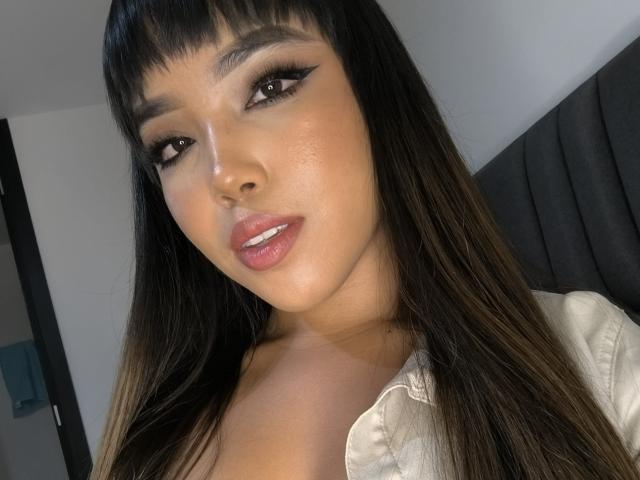 Heyly has long black hair, heavy eyeliner, and glossy pink lips, looking seductively at the camera with a slight smile.