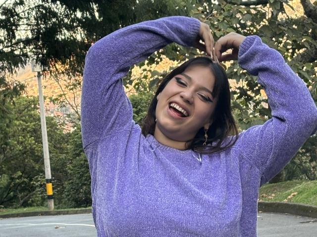 MillyJohnson with medium tits, wearing a purple sweater, smiling with arms raised, hands touching head, brown hair, green eyes, outdoors, trees in background.