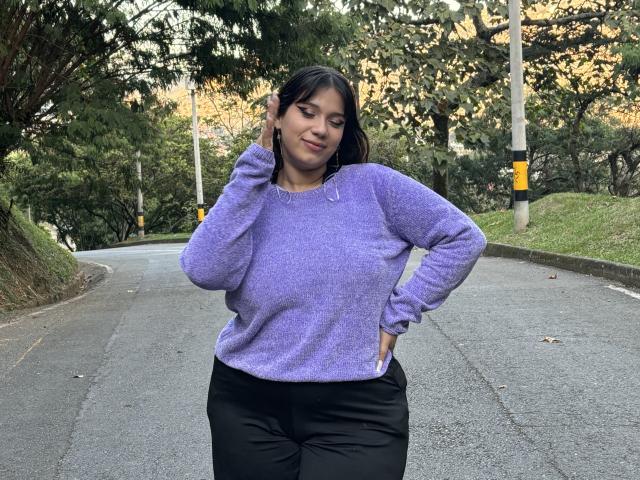 Milly Johnson, con cabello oscuro, un suéter morado y pantalones negros, está de pie en una calle, con la mano en la oreja, sonriendo, mostrando su trasero curvilíneo y sus pequeños senos. Exterior, de día..