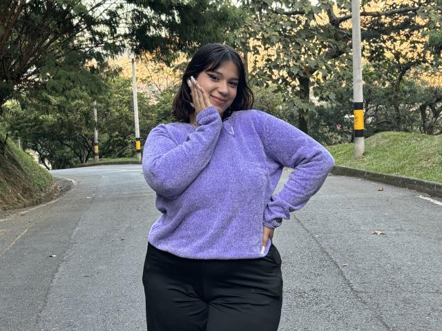 Milly Johnson, con piel de tono marrón medio, cabello oscuro y ondulado, vestida con una sudadera morada y pantalones negros, con la mano en la mejilla, sonriendo, de pie en una carretera de asfalto curva..