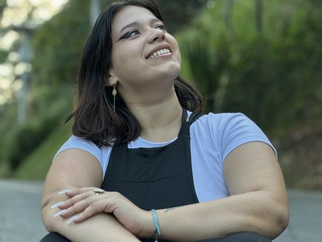 MillyJohnson with medium tits, smiling, arms crossed, wearing black apron over white shirt, dark brown hair, blue earrings, bracelet, green foliage background, daytime, natural light.