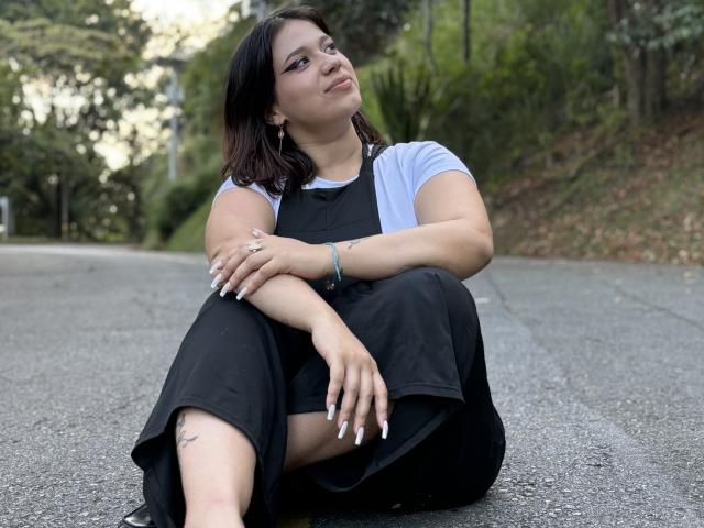 MillyJohnson sitting on a road, black dress over white shirt, crossed arms, looking up, short brown hair, white nails, tattoo on right thigh, green bracelet, relaxed.