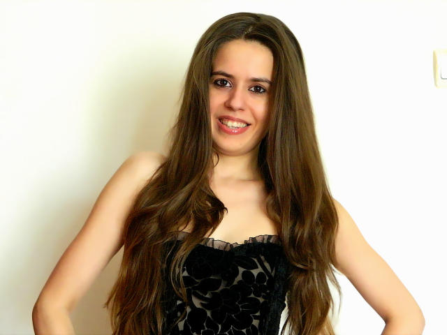 LonelyAngel69 with long brown hair, smiling, wearing a black floral strapless dress, hands on hips, showing her tits, against a plain white background.