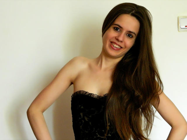 LonelyAngel69, com cabelos castanhos longos, sorrindo, vestindo uma blusa preta de renda sem alças, com as mãos nos quadris, mostrando os seios e o bumbum de forma discreta..