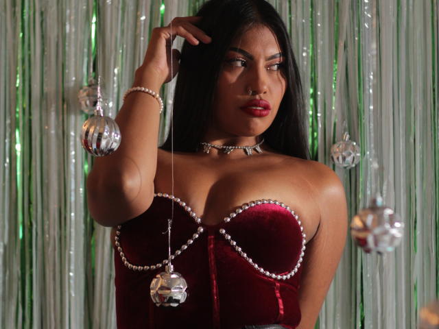 KimJohnsson con capelli neri, un corpetto di velluto rosso con bordi di perle, grandi seni, pantaloni neri, che tiene una campanella d'argento, sorridente, in piedi accanto a una scatola regalo con un nastro verde..