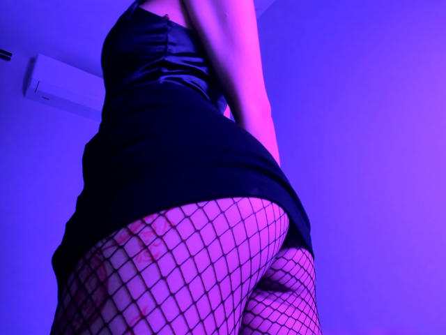 MishelBlessing's ass in black fishnet stockings, wearing a short black dress, under purple and blue lighting. Her skin is visible through the fishnets, showcasing her curves.