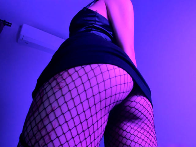 MishelBlessing's ass in fishnet stockings, wearing a black dress, shot from a low angle with purple and blue lighting, highlighting her butt and upper thighs.