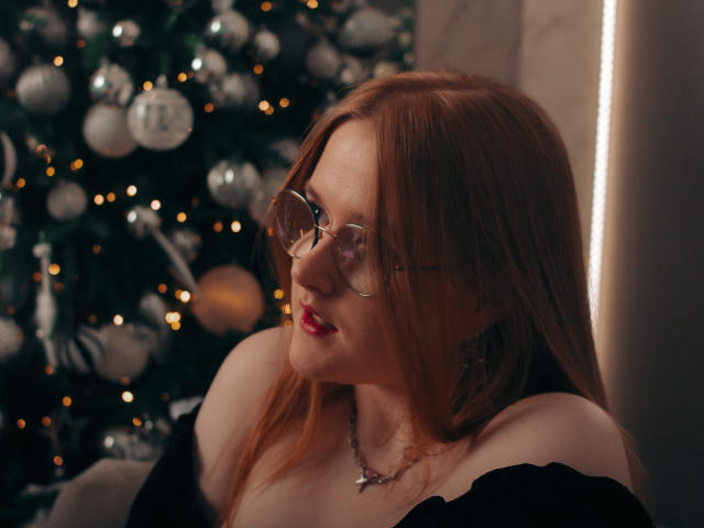 BonnieBell with red hair, glasses, and red lipstick, wearing a black off-shoulder top, looking to the side, showing her tits, next to a decorated Christmas tree.