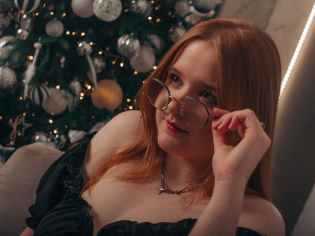 BonnieBell with red hair, glasses, and black off-shoulder top, touching her glasses, showing her tits, in front of a decorated Christmas tree, smiling.