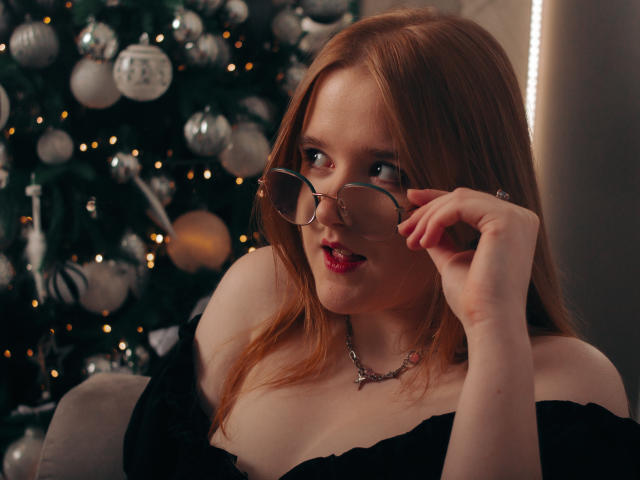 BonnieBell with red hair, wearing glasses, black off-shoulder top, looking up with a slight smile, touching glasses, showing tits, necklace, Christmas tree with silver ornaments in background.