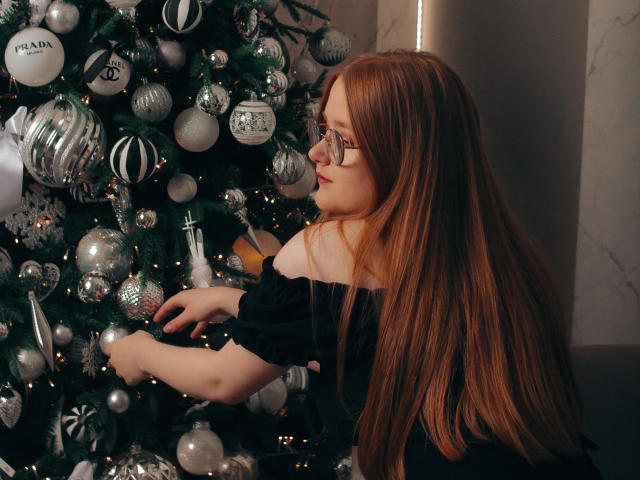 BonnieBell with long red hair and glasses, wearing a black off-shoulder top, decorating a silver and white Christmas tree with shiny ornaments. Her tits are partially visible.