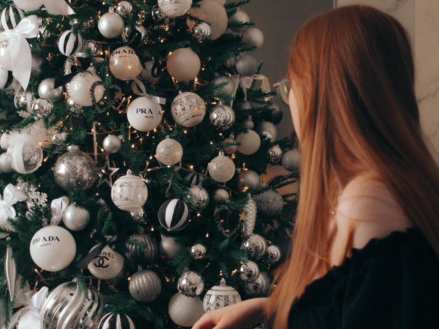 BonnieBell with red hair stands in front of a silver and white Christmas tree adorned with Prada ornaments, glittering lights, and shiny baubles, wearing a black off-shoulder top.