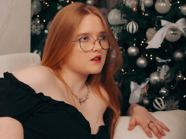BonnieBell with red hair, glasses, and red lipstick, wearing a black off-shoulder top, necklace, and ring, sitting by a decorated Christmas tree. Her tits are partially visible.