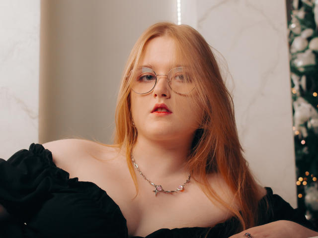 BonnieBell has red hair, glasses, and red lips. She's wearing an off-shoulder black top and a silver necklace. Her tits are partially visible. She looks serious.
