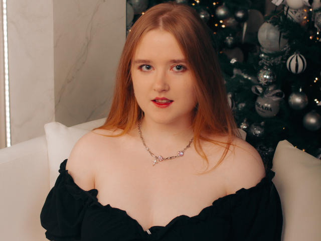 BonnieBell has red hair, pale skin, blue eyes, and wears a black off-shoulder top with a delicate necklace. She's sitting by a Christmas tree with silver ornaments.
