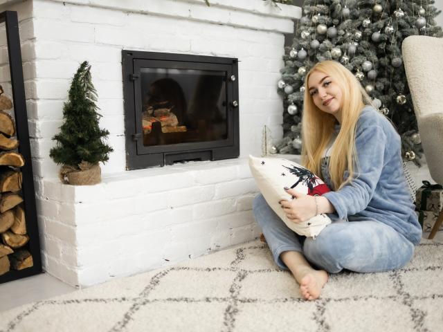 BeatriceFly, blond, in blauen Pyjamas, sitzt auf einem weißen Teppich, hält ein Weihnachtskissen, vor einem schwarzen Kamin, mit einem kleinen Baum und einem silbernen Weihnachtsbaum im Hintergrund. Barfuß, lächelnd, niedlich..