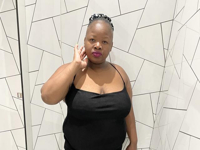 BustySasha with dark skin, purple lipstick, black tank top showing her tits, holding phone to ear, short black hair with white pattern, standing in white geometric tiled room.