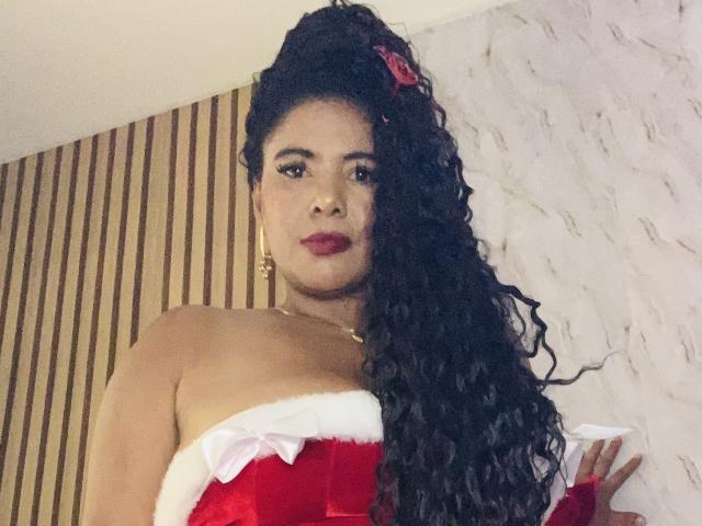 Sibeli with long curly black hair, red lipstick, wearing a red velvet strapless dress with white fur trim, looking down, gold earrings, marble wall background, confident expression.