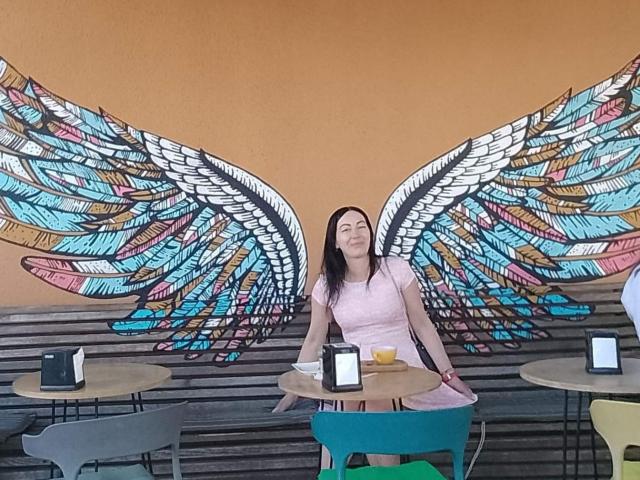 MirandaHott69, sitting at a table, wearing a pink top, smiling, with large colorful angel wings painted on the wall behind her, in a café with assorted chairs.