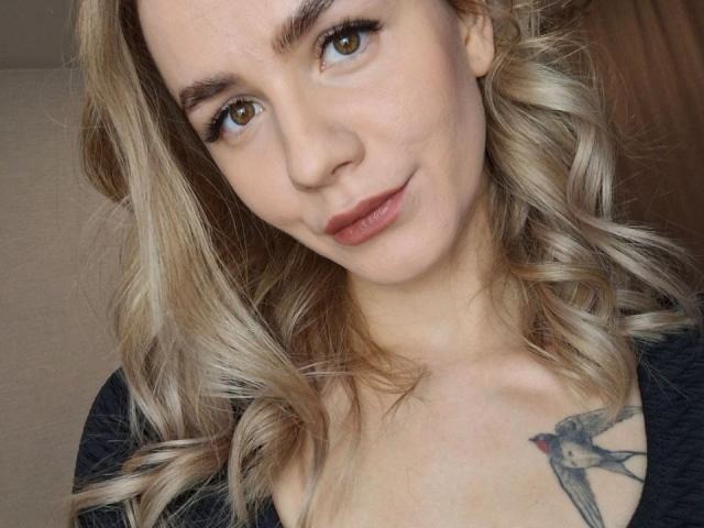 InfiniteDesire has blonde, wavy hair, brown eyes, and a small tattoo of a bird on her right chest. She's wearing a black top and has a slight smile.