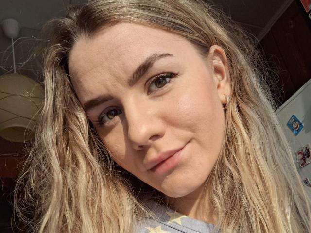 InfiniteDesire has blonde, wavy hair, green eyes, and a subtle smile. She's wearing a light-colored shirt with a yellow pattern. Her small nose and light makeup are visible.