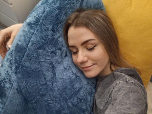InfiniteDesire with closed eyes, lying on a blue fuzzy pillow, smiling slightly. She has long brown hair, a gray shirt, and is next to a yellow pillow.