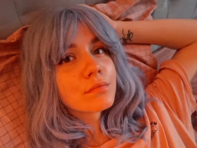 InfiniteDesire with purple-blue hair, pouty lips, and a tattoo on her forearm, lying on an orange bed, wearing an orange shirt, with a warm orange light.