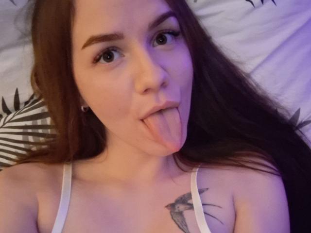 InfiniteDesire with long brown hair, sticking out her tongue, wearing a white tank top, showing a bird tattoo on her chest, with green eyes, and light skin.