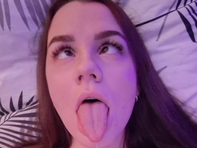 InfiniteDesire's face with long brown hair, green eyes, and tongue sticking out, looking up with a slight purple glow, wearing small earrings, and looking playful.