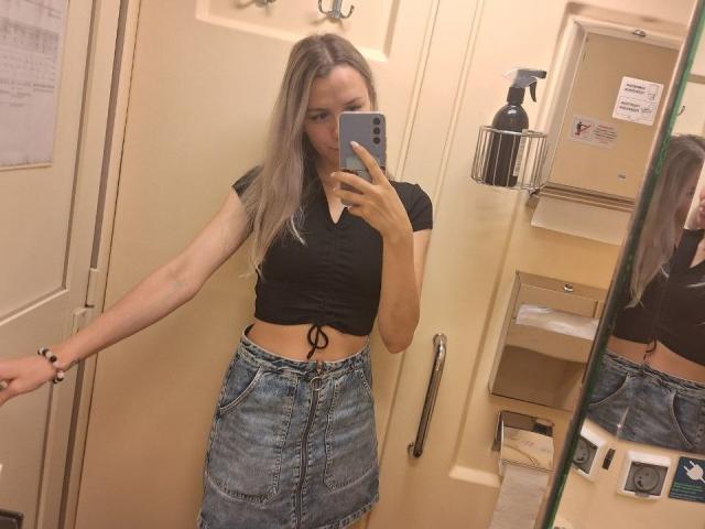 InfiniteDesire with long blonde hair, black crop top, tied at waist, blue denim skirt, holding phone in mirror selfie, right arm extended, mirror reflection, bathroom.