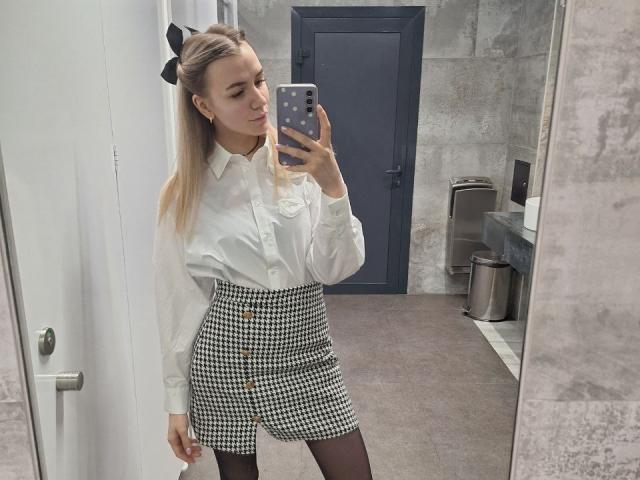 InfiniteDesire taking a mirror selfie, wearing a white shirt, black and white checkered skirt, black stockings, and a black bow in her blonde ponytail. Her phone has a polka-dot case.