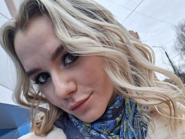 InfiniteDesire has wavy blonde hair, blue eyes, and a cute smile. She's wearing a blue and yellow patterned scarf. Her nose is small and her skin is fair.