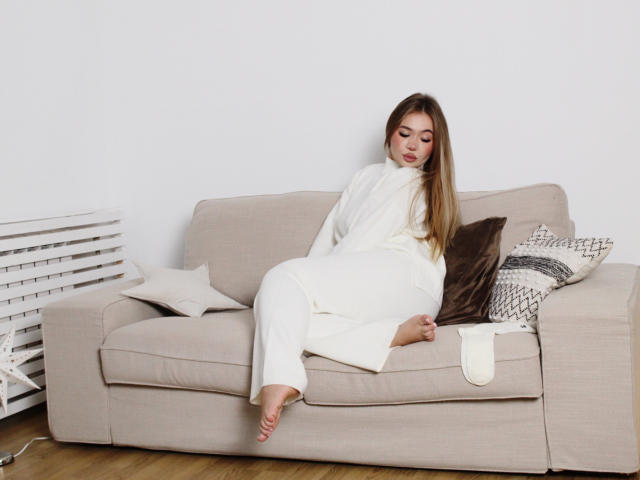 Stacy Yellig, con lunghi capelli castani, indossa una maglia a collo alto bianca e pantaloni, è seduta su un divano beige, con gli occhi chiusi e i piedi nudi. Ci sono cuscini marroni e bianchi, una parete bianca e un pavimento in legno..