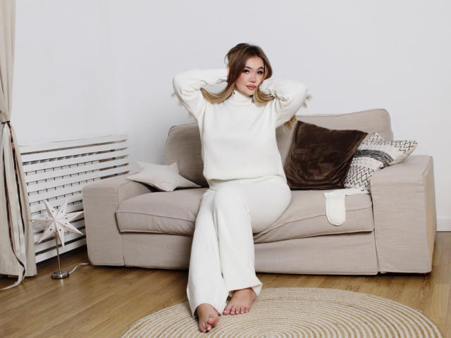 Stacy Yellig está sentada en un sofá beige, vestida con un cuello alto blanco y pantalones blancos, con las manos en el pelo, los pies descalzos sobre una alfombra redonda, y parece relajada..