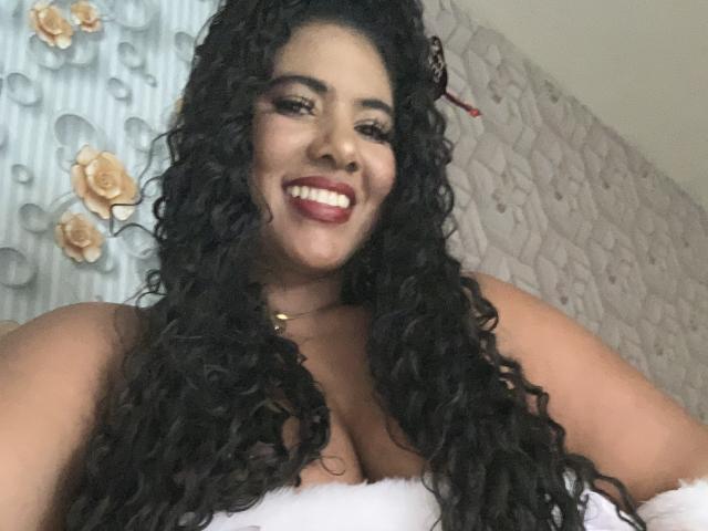 Sibeli with curly black hair, smiling, showing her tits, wearing a white fluffy top, red lipstick, gold necklace, and a red hair accessory in a selfie.