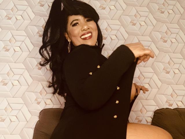 Sibeli with dark hair, smiling, wearing a black button-up dress, showing her tits, arms raised, sitting on a brown couch, gold hoop earrings, and a cheerful expression.