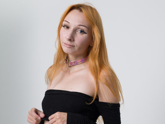 HannaCool with long red hair, black off-shoulder top, nose ring, and pink plaid choker, holding her tits, smiling slightly.