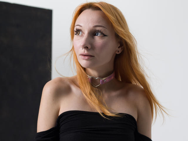 HannaCool with orange hair, black off-shoulder top, pink collar with metal ring, looking to the side, fair skin, subtle makeup, small nose ring, and relaxed expression.