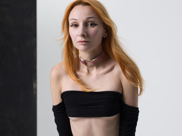 HannaCool with orange hair, black strapless top, small boobs, pink choker, and eyeliner, standing with arms behind her back, looking at the camera.